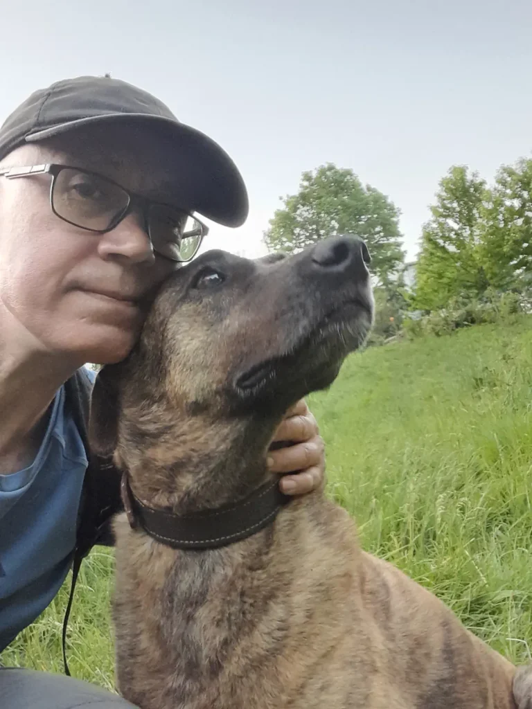 A close-up selfie of a man [Özgür Nevres] gently holding Lady, a brown brindle rescue dog, as she leans against him in a grassy sanctuary area, looking upward with calm eyes.