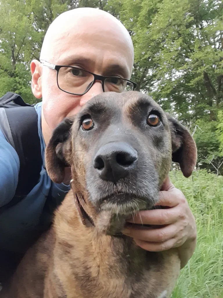 A close-up selfie of a man [Özgür Nevres] holding Lady the rescue dog, her expressive eyes looking toward the camera as they sit together on the grass with trees in the background.