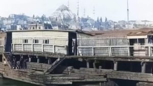 Colorized frame from the oldest video of istanbul [1897] showing the old Galata Bridge over the Golden Horn, with wooden structures, boats on the water, pedestrians on the bridge, and the Historic Peninsula with domes and minarets in the background